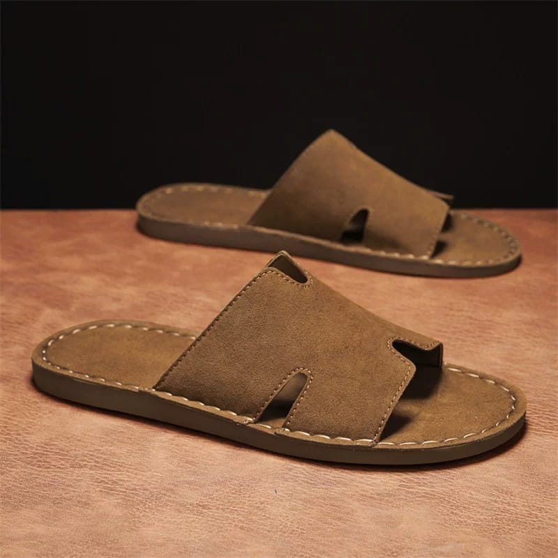 Pair of brown suede slide sandals with H-shaped cutout straps on a warm leather surface with a dark backdrop