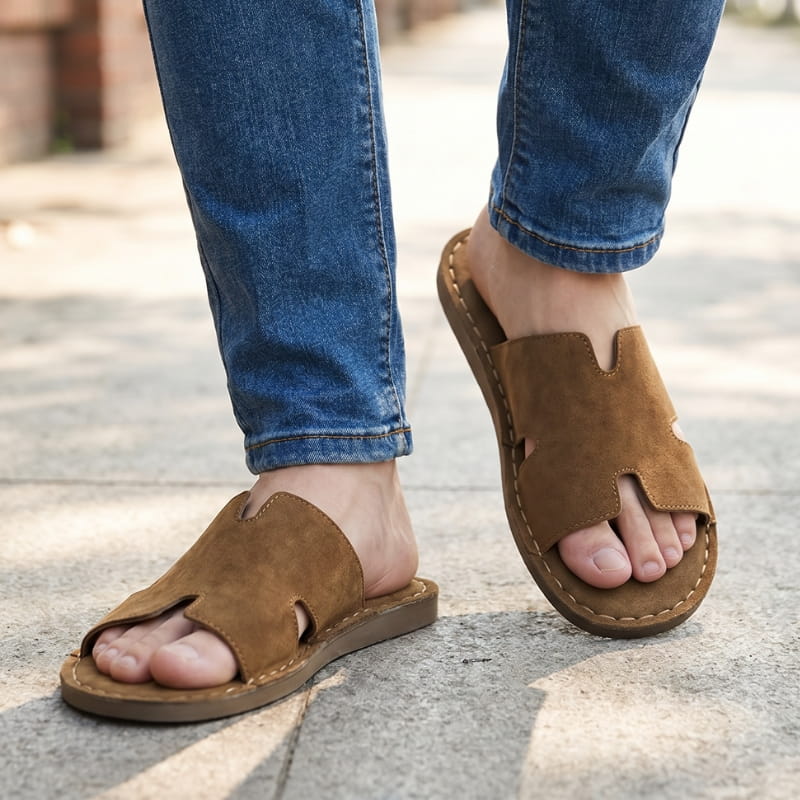 Legs in cuffed jeans wearing brown suede slide sandals, walking on outdoor pavement in sunlight