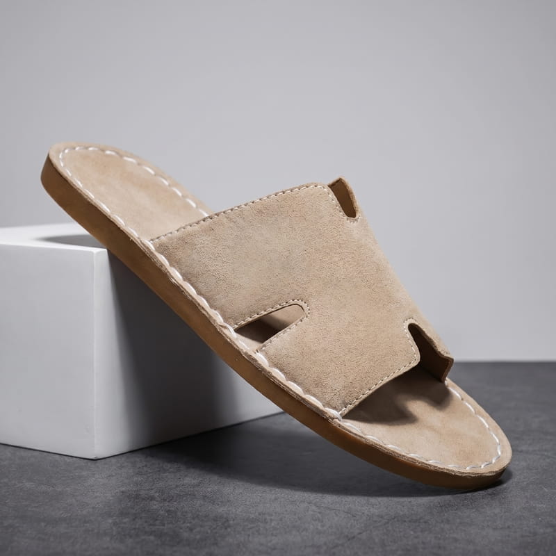 Khaki suede slide sandal with H-shaped cutout strap and hand-stitched trim propped diagonally against a white geometric block