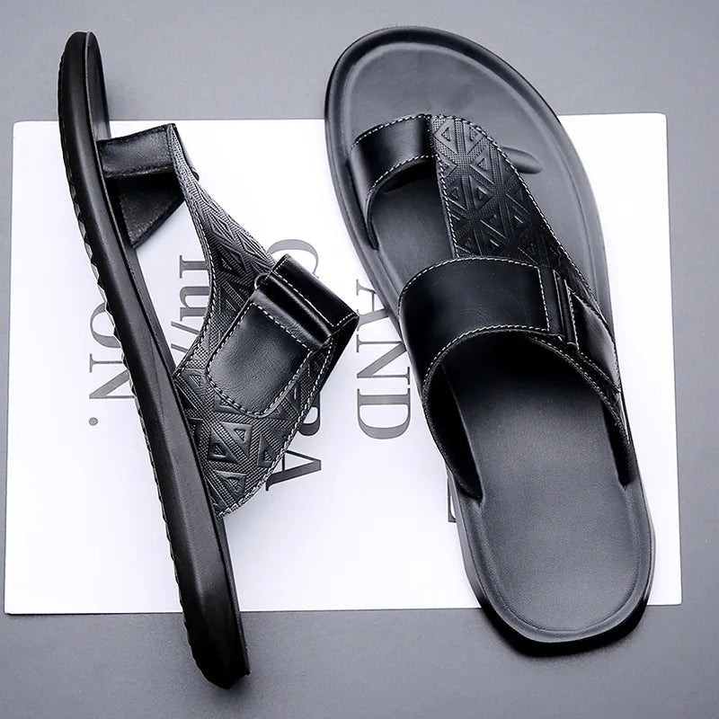 Tilted overhead view of a pair of black leather sandals with geometric-textured toe-loop straps, resting on branded paper packaging.