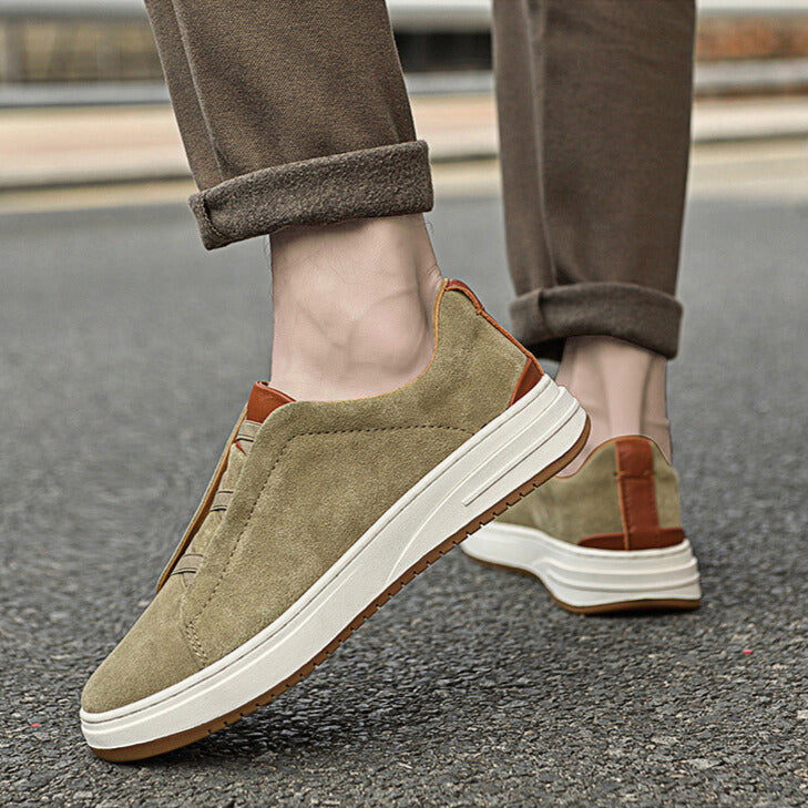 Close-up of feet walking on pavement wearing olive suede Dominic slip-on sneakers with cuffed brown trousers