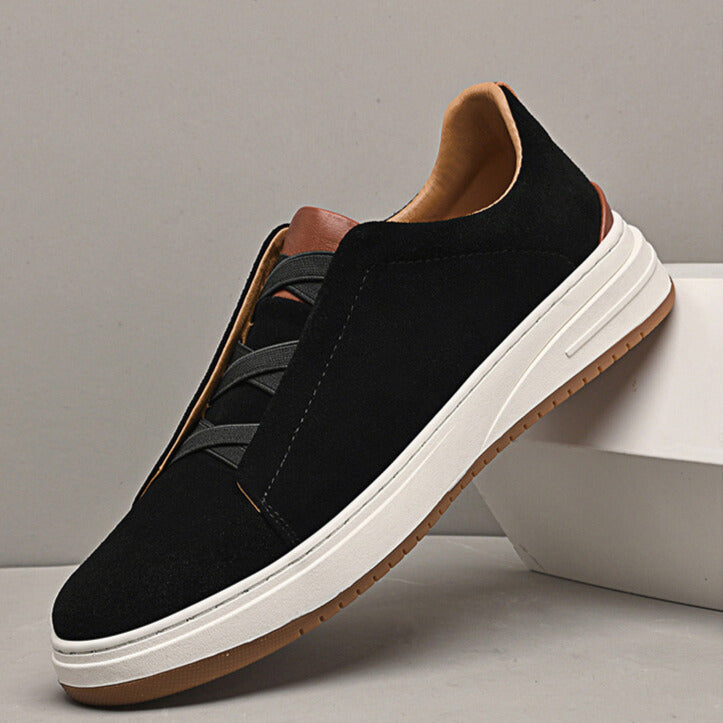 Single black suede Dominic slip-on sneaker with elastic gore lacing and tan trim, leaning against a white box