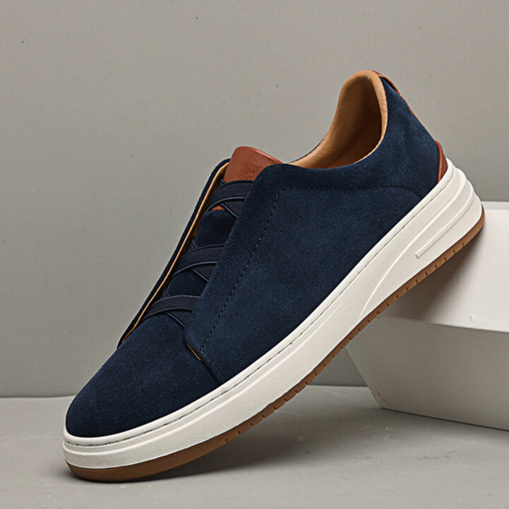 Single navy blue suede Dominic slip-on sneaker with elastic gore lacing and tan trim, leaning against a white block