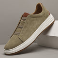 Single olive suede Dominic slip-on sneaker with elastic gore lacing and white sole, leaning against a white box