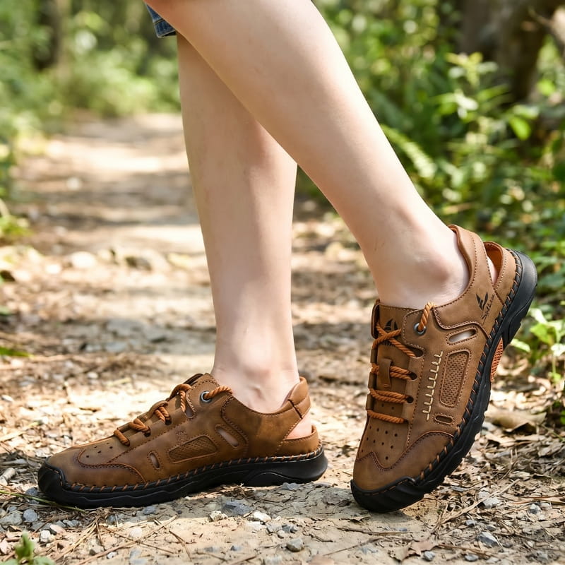 Bare ankles wearing brown nubuck fisherman sandals, walking on a gravel path through a wooded area