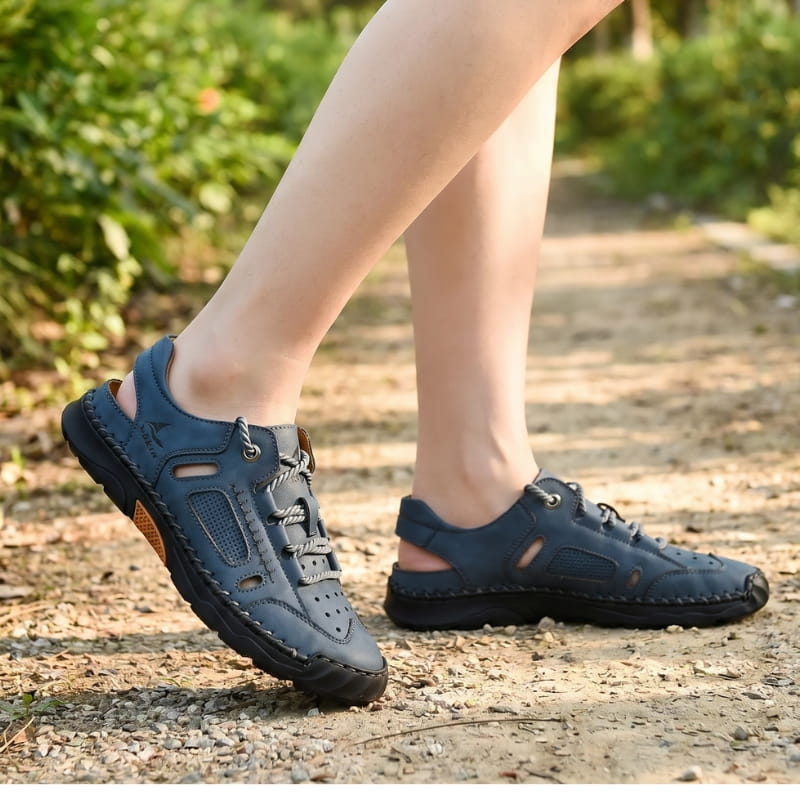 Bare ankles wearing blue nubuck fisherman sandals, walking on a dirt path lined with greenery