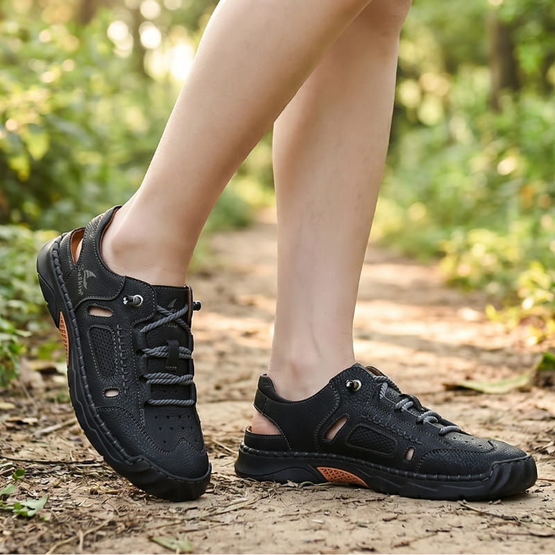 Bare ankles wearing black leather fisherman sandals with orange trim, walking on a wooded dirt path