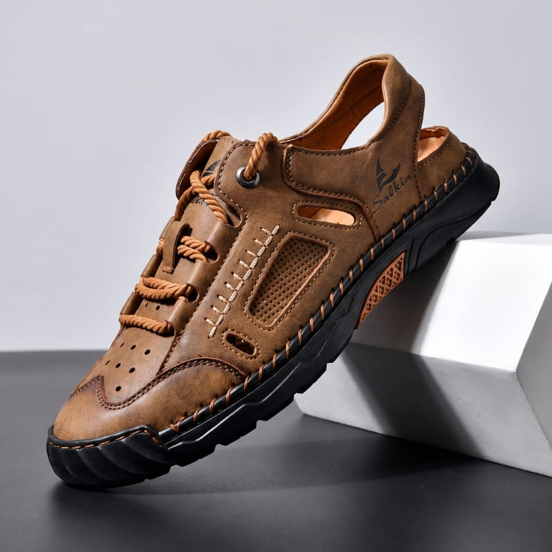 Brown nubuck leather fisherman sandal with braided laces and perforated toe cap propped diagonally against a white geometric block