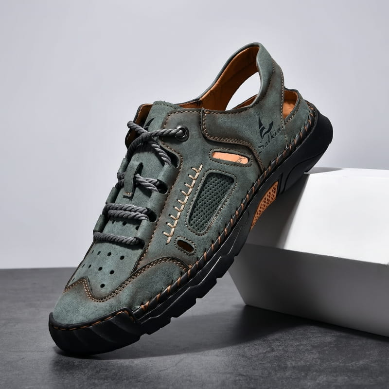 Green nubuck leather fisherman sandal with rope-style laces and perforated panels propped diagonally against a white geometric block