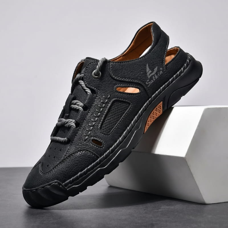 Black leather fisherman sandal with rope-style laces and orange trim accents propped diagonally against a white geometric block