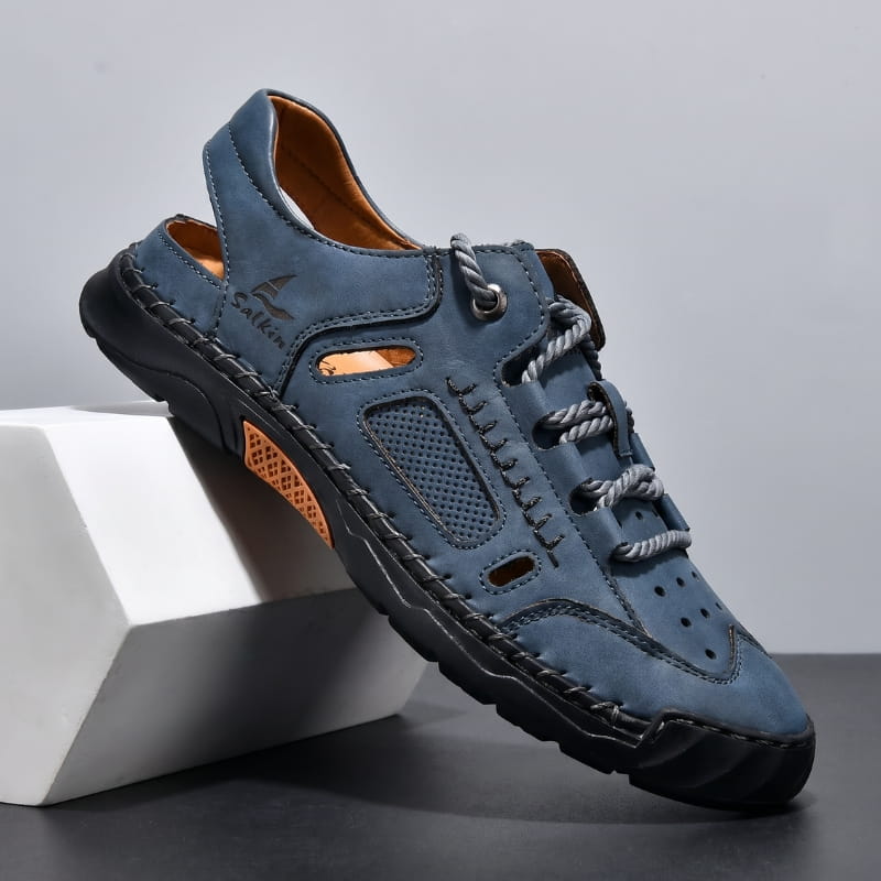 Blue nubuck leather fisherman sandal with orange lining, perforated side panels, and toggle laces propped diagonally against a white geometric block