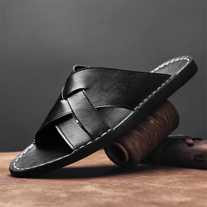 Black woven leather cross-strap sandal with a cushioned flat sole, styled on a wooden surface beside a rolled leather strip.