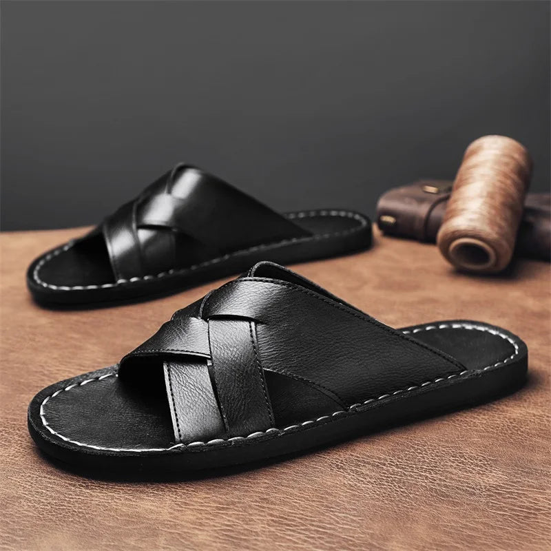 Pair of black woven leather cross-strap sandals resting on a wooden surface beside a rolled leather strip.