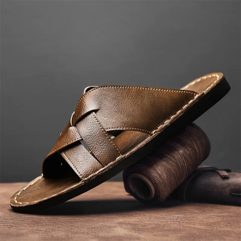 Brown woven leather cross-strap sandal with a cushioned flat sole, styled on a wooden surface beside a rolled leather strip.