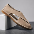Khaki suede slip-on sandal with cross-wrap cutout design propped diagonally against a white geometric block