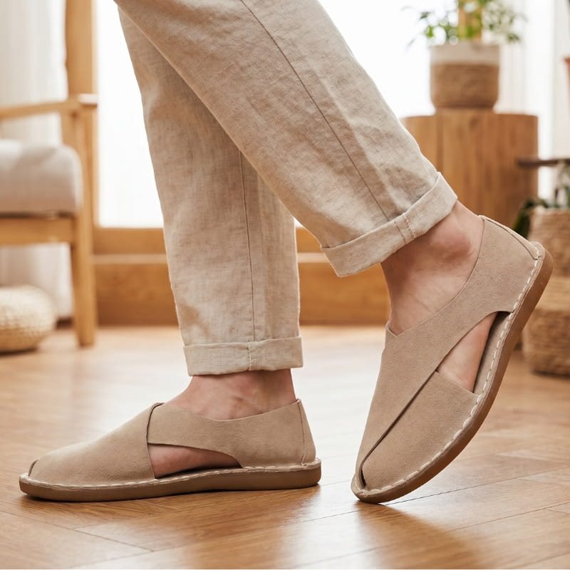 Legs in cuffed beige linen trousers wearing khaki suede slip-on sandals, standing on a wooden floor indoors