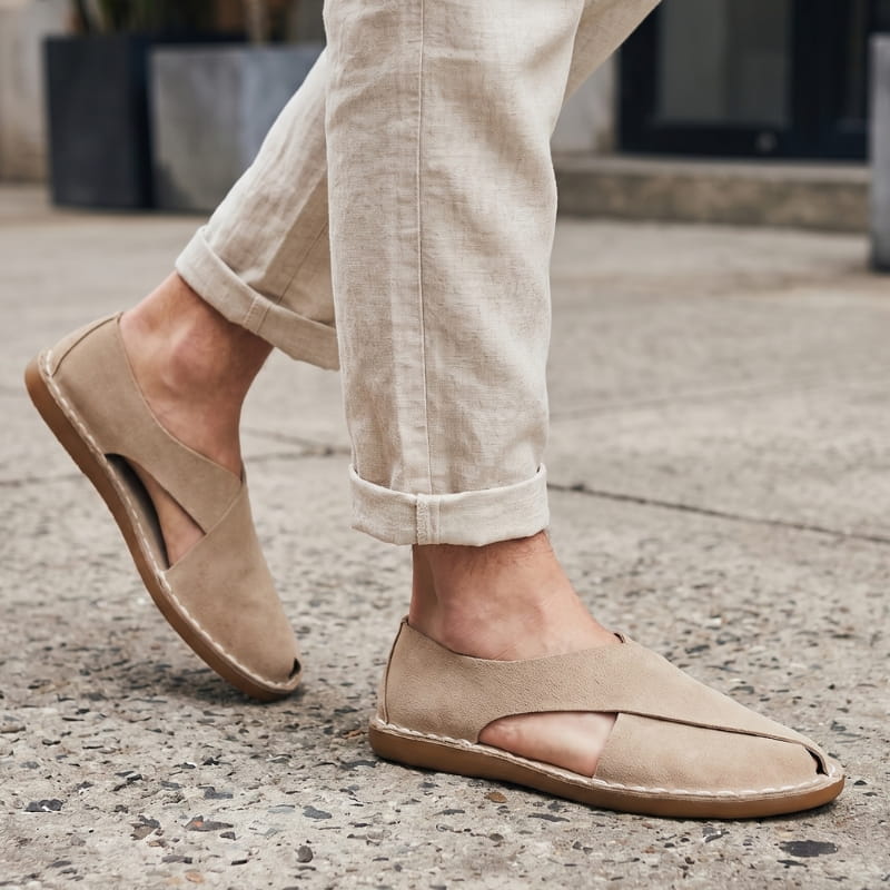 Legs in cuffed beige linen trousers wearing khaki suede slip-on sandals, walking on an outdoor concrete sidewalk