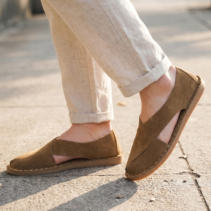 Legs in cuffed beige linen trousers wearing brown suede slip-on sandals, walking on a sunlit outdoor pavement