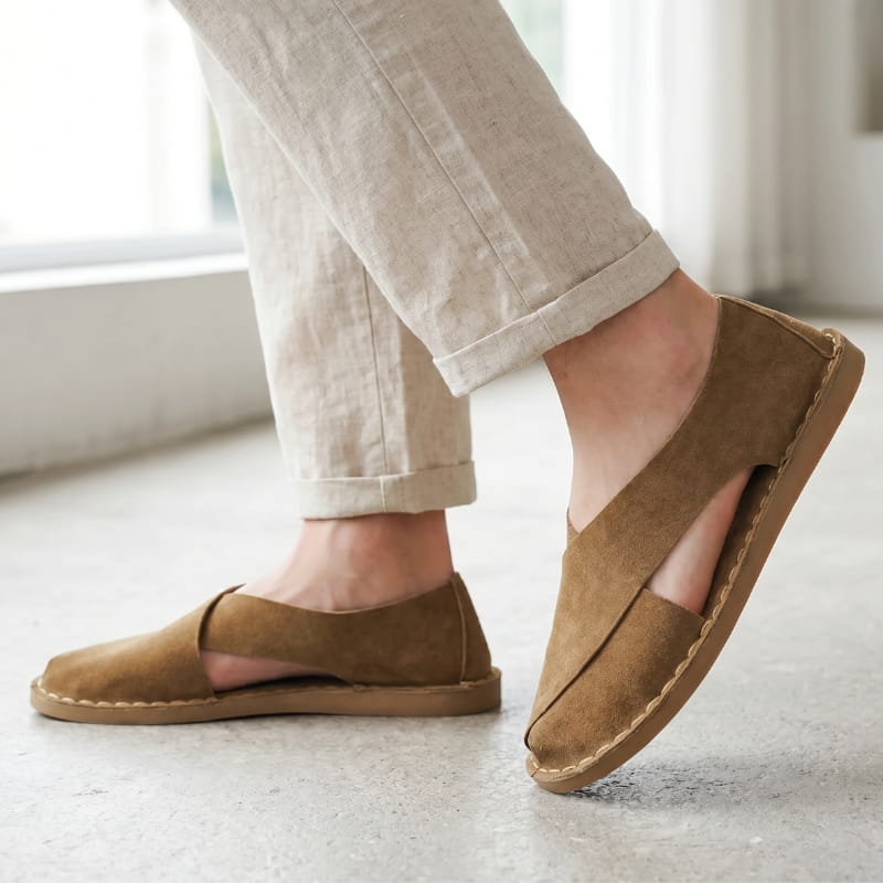 Legs in cuffed beige linen trousers wearing brown suede slip-on sandals, standing on a concrete floor near a bright window