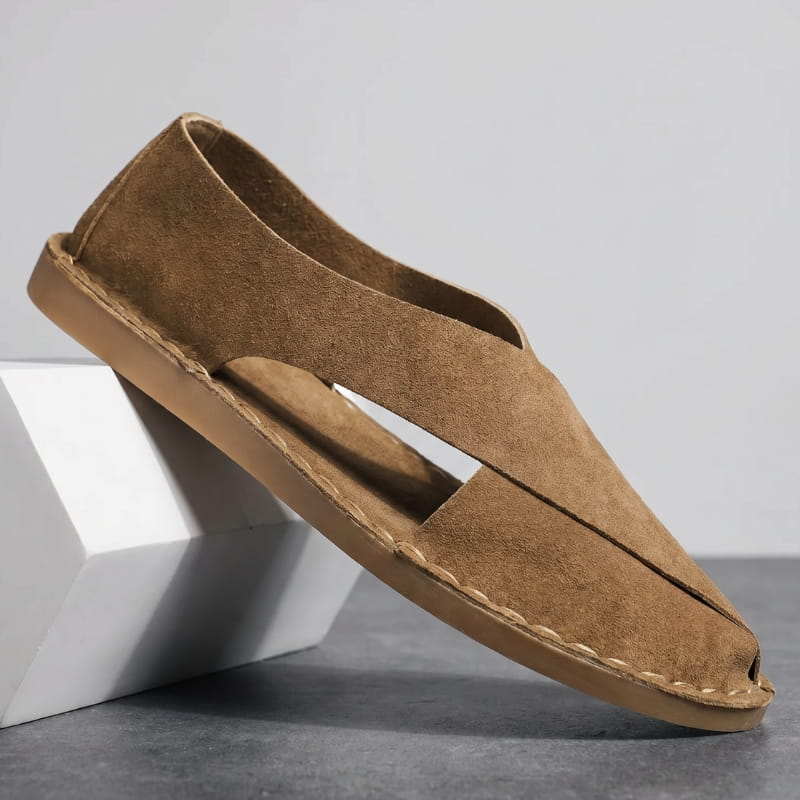 Brown suede slip-on sandal with cross-wrap cutout design propped diagonally against a white geometric block