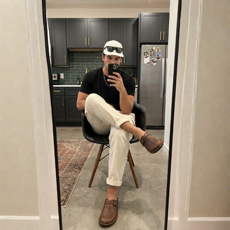 Mirror selfie of a man in a cap and black t-shirt seated on a stool wearing brown leather buckle mules in a kitchen