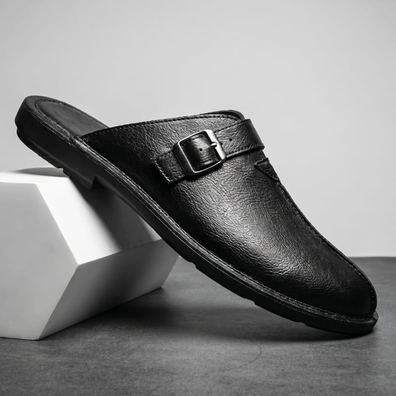Black leather mule with polished buckle strap propped diagonally against a white geometric block in a studio setting