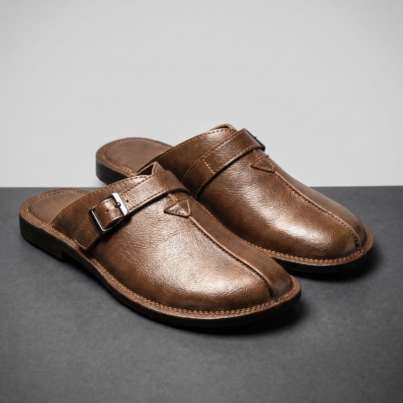 Pair of brown leather mules with polished buckle straps on a gray backdrop