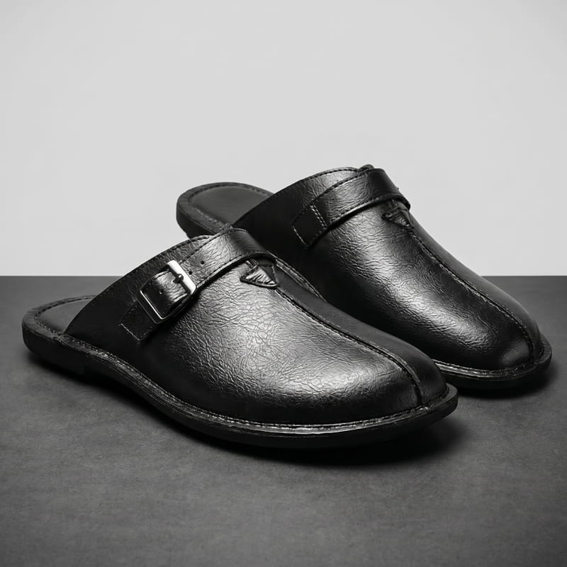 Pair of black leather mules with polished buckle straps on a gray backdrop