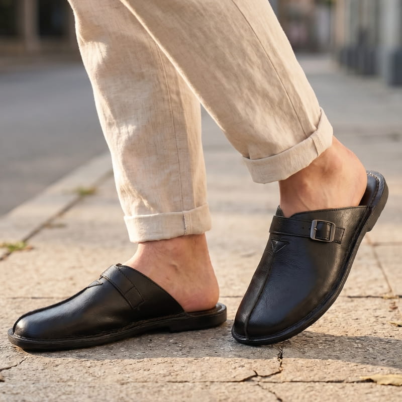 Legs in cuffed beige linen trousers wearing black leather buckle mules, walking on a cobblestone street