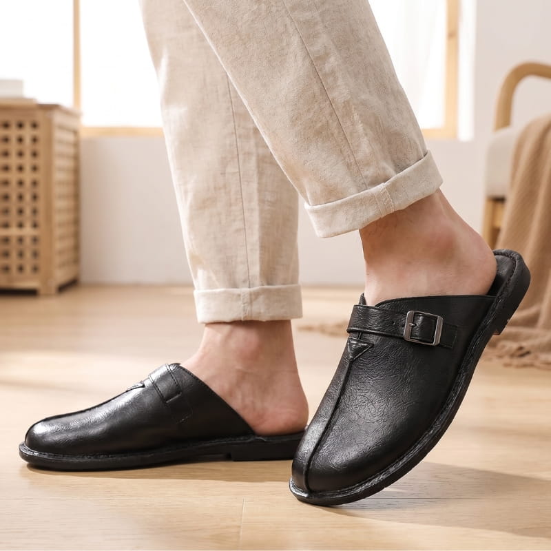 Legs in cuffed beige linen trousers wearing black leather buckle mules, standing on a wooden floor near a window