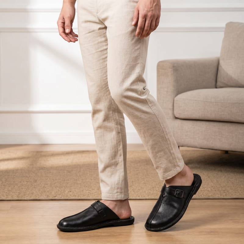 Legs in beige trousers wearing black leather buckle mules, standing in a living room near a sofa