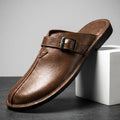 Brown leather mule with polished buckle strap propped diagonally against a white geometric block in a studio setting