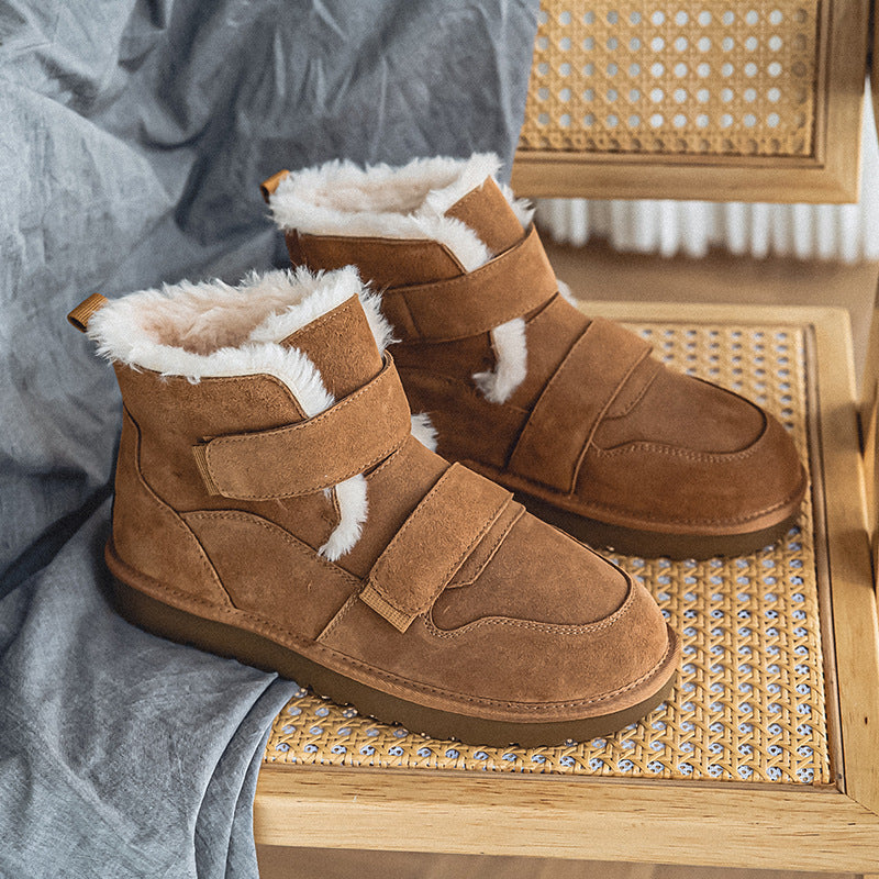 Pair of cognac suede fleece-lined winter boots with velcro straps, resting on a woven cane chair with a gray fabric drape.