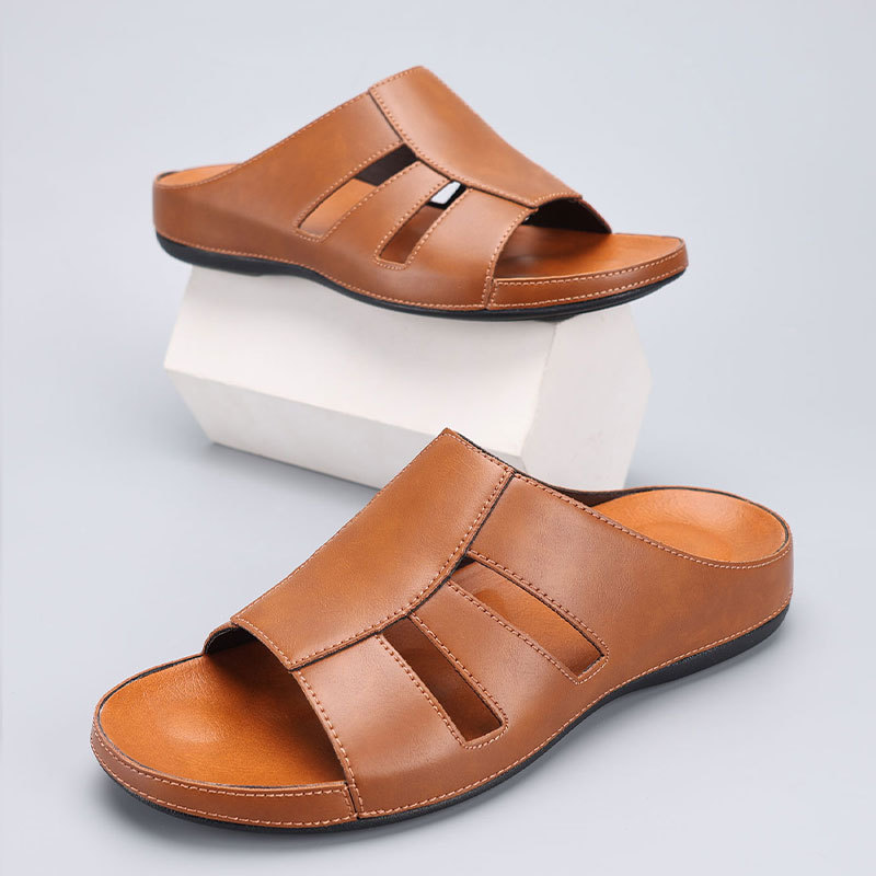 Pair of tan leather slip-on sandals with cutout straps, one shoe propped on a white display block above the other.