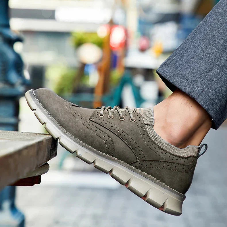 A gray suede sneaker rests on a stone ledge, with cuffed gray trousers and a blurred urban street in the background.