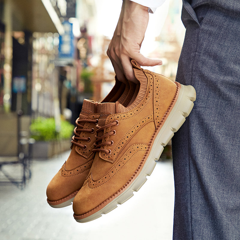 A hand holds tan suede brogue-style sneakers by the heel, with a blurred city street in the background.