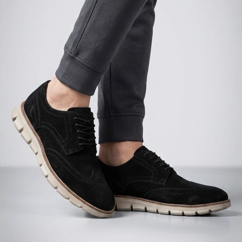 Close angled view of a person's feet wearing black suede sneakers and gray joggers in a studio setting.
