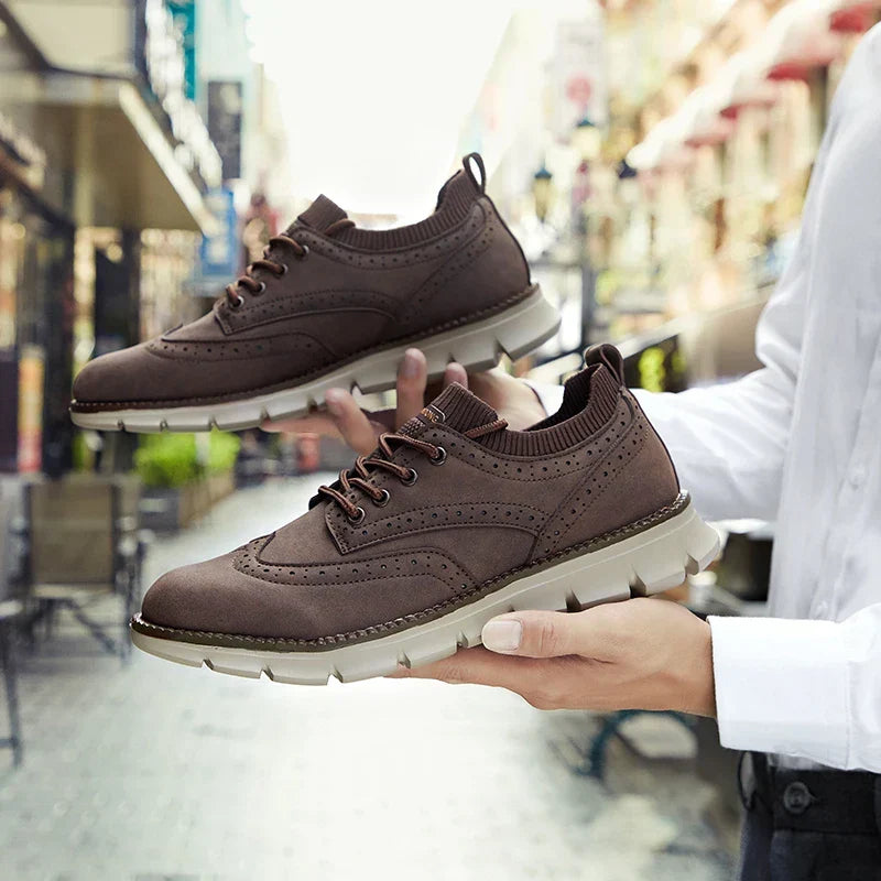 A hand holds dark brown suede brogue-style sneakers, with a blurred shopping street in the background.