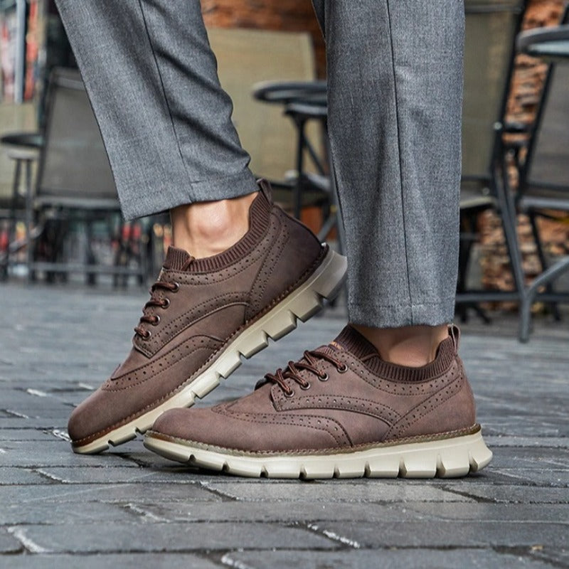 A person walking on a cobblestone street wearing dark brown suede sneakers and gray trousers.