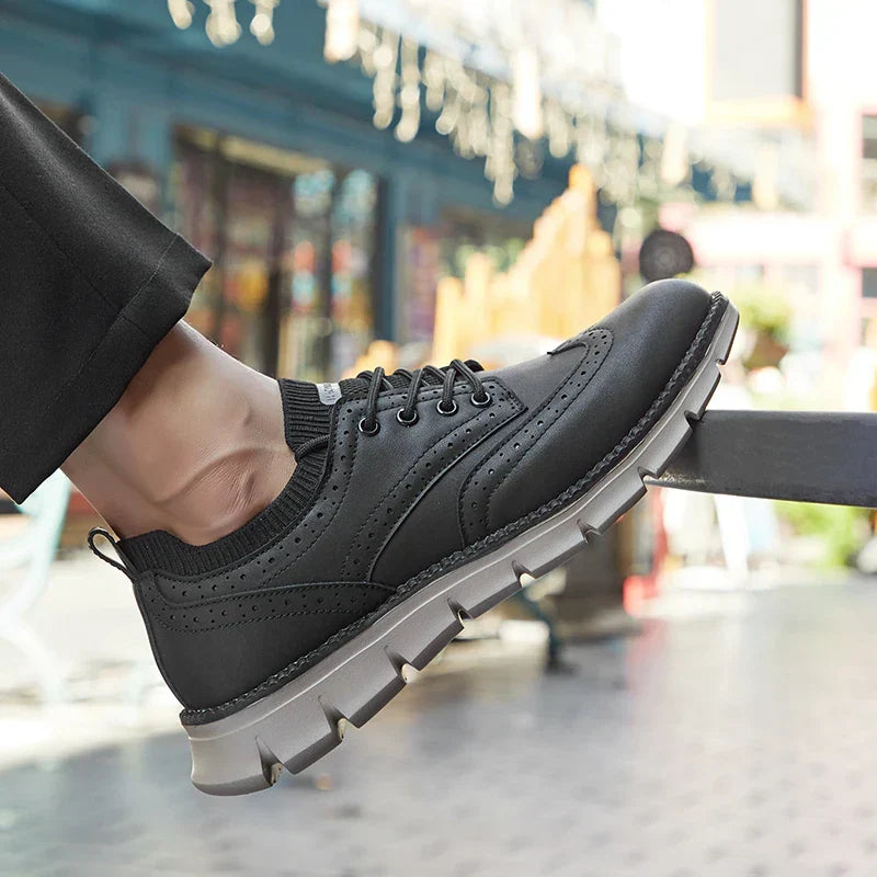 Close-up angled shot of a person's foot wearing a black brogue-style sneaker with dark trousers, blurred street scene in the background.