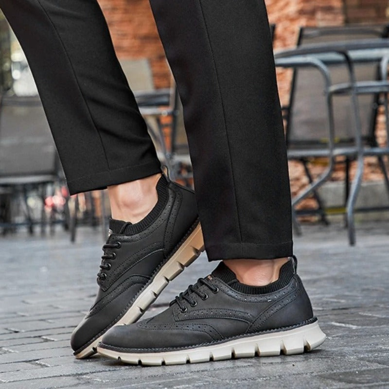 A person crossing their legs while standing on cobblestone, wearing black brogue-style sneakers and black trousers.