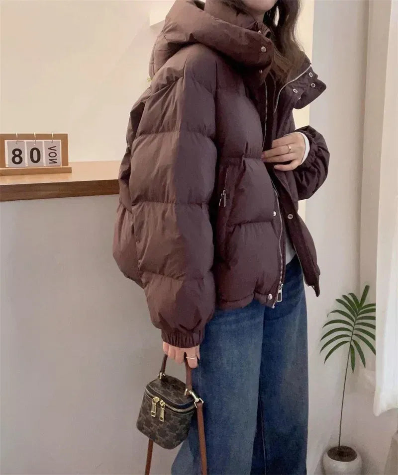 Dark brown quilted puffer jacket with a high funnel neck, attached hood, and relaxed fit worn over blue jeans.