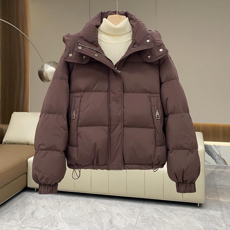 Dark brown hooded puffer jacket with quilted insulation, high funnel neck, and zip pockets, layered over a cream turtleneck.