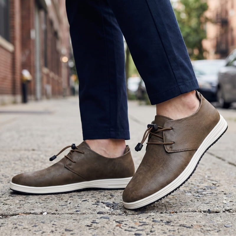 Man wearing brown leather lace-up sneakers with navy trousers, walking on a city sidewalk
