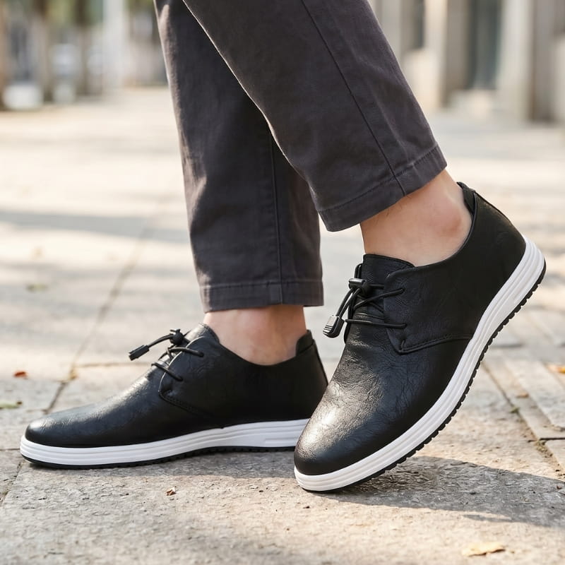 Man wearing black leather lace-up sneakers with gray trousers, walking on a paved outdoor path