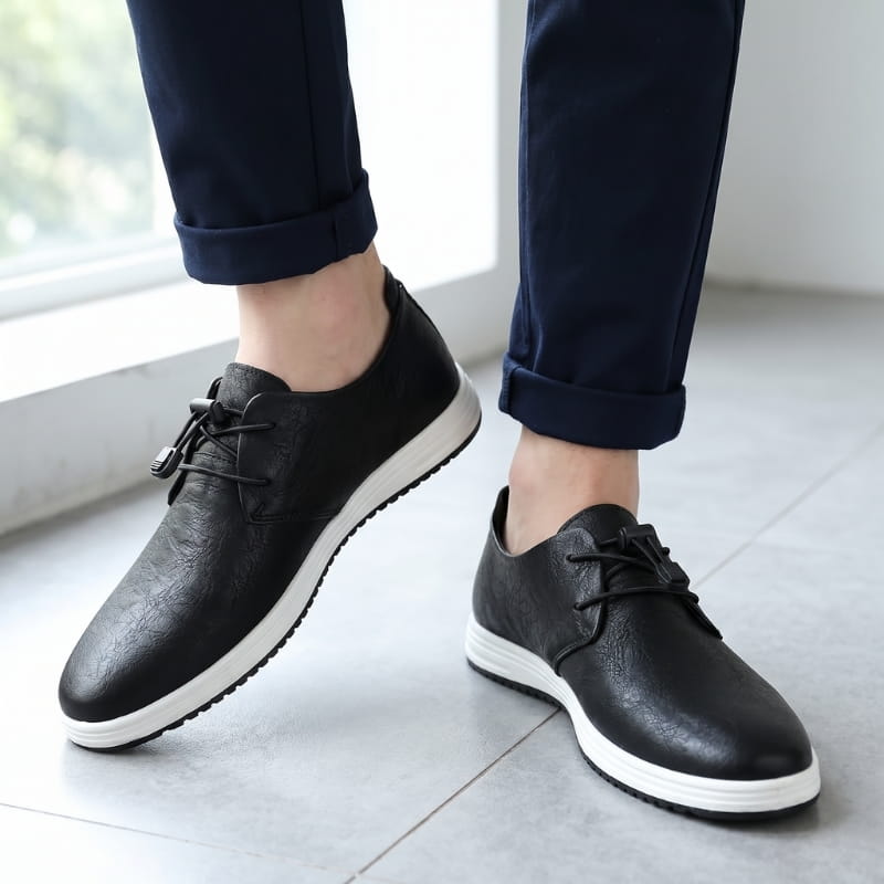 Man wearing black leather lace-up sneakers with cuffed navy trousers, standing indoors by a bright window