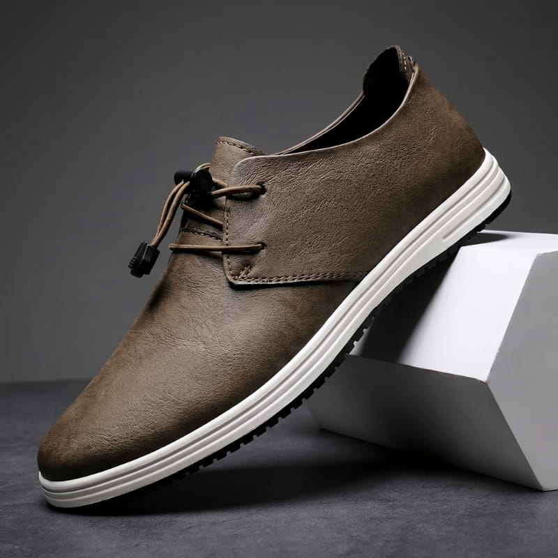 Brown leather lace-up sneaker with white sole propped diagonally against a white geometric block in a studio setting