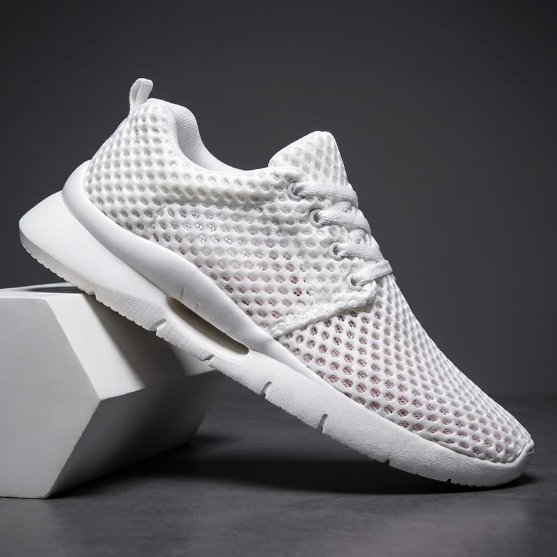 White mesh sneaker with white cushioned sole propped diagonally against a white geometric block in a studio setting
