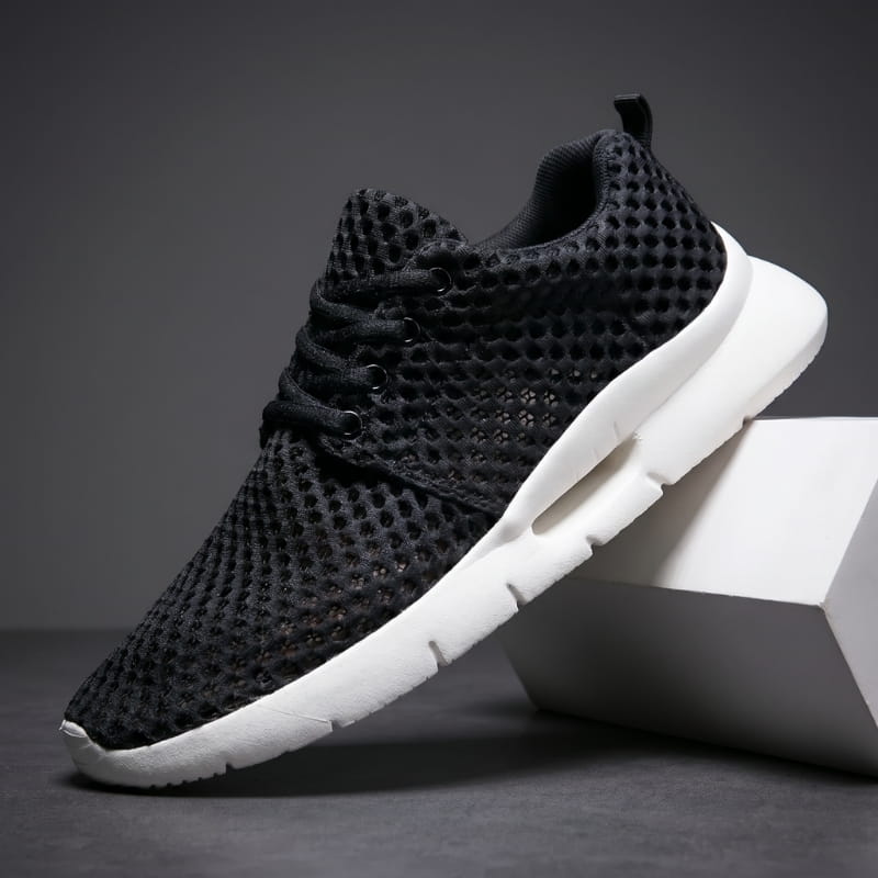 Black mesh sneaker with white cushioned sole propped diagonally against a white geometric block in a studio setting