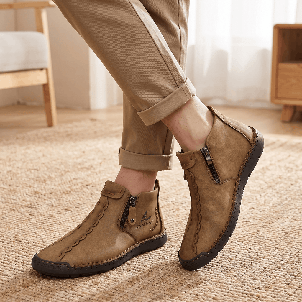 Close-up of crossed legs wearing tan trousers and brown leather Javier high-top zip boots standing on a jute rug near a sunlit window